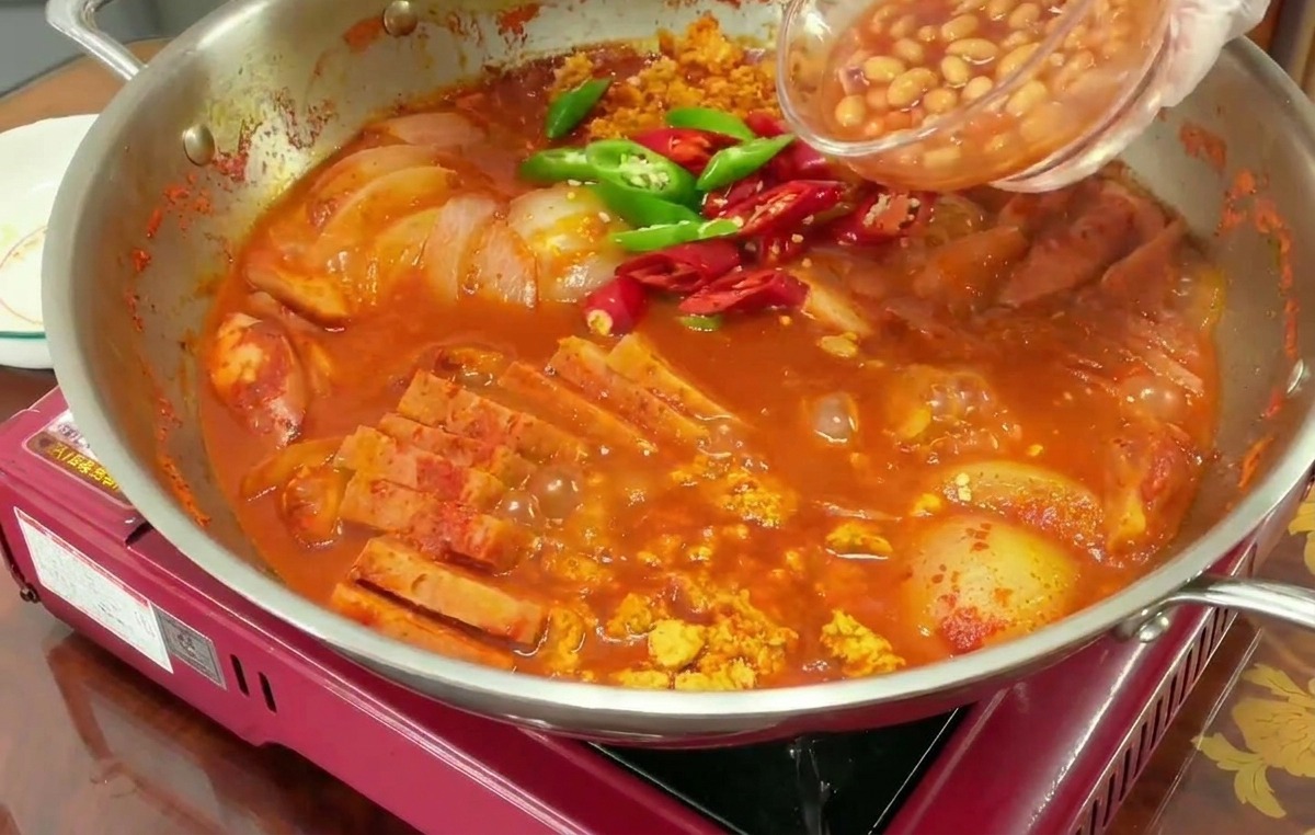 A hand pouring baked beans from a small glass bowl into a bubbling red pot of stew already containing sliced green and red chili peppers.