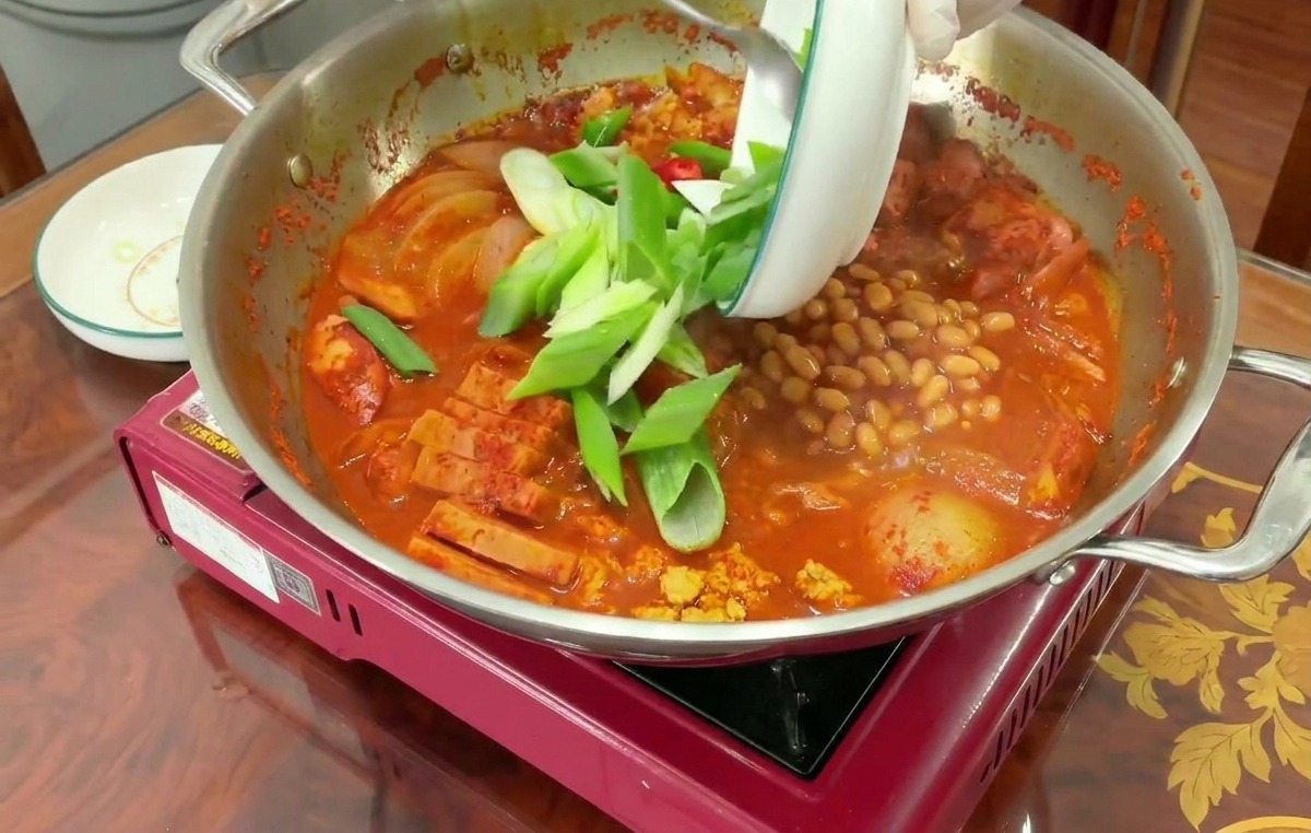 Sliced green leeks being poured from a white ceramic bowl onto the surface of a boiling red Army Base Stew.