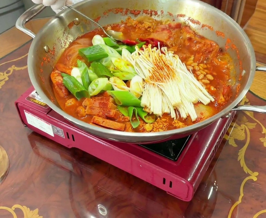 Gloved hands using a spoon to stir a bubbling pot of Budae Jjigae, mixing the meat, vegetables, and seasoned red broth together.