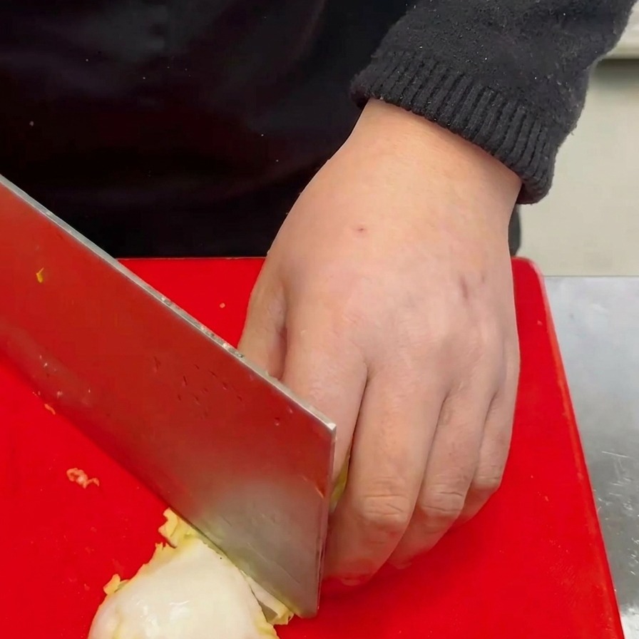 A hand using a large cleaver to chop a head of white baby cabbage into chunky pieces on a bright red cutting board.