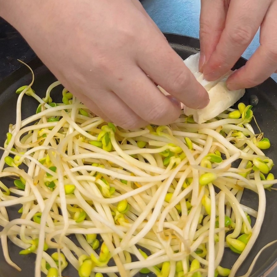 Hands carefully placing thick slices of white tofu over a bed of bean sprouts in a shallow black cooking pot.