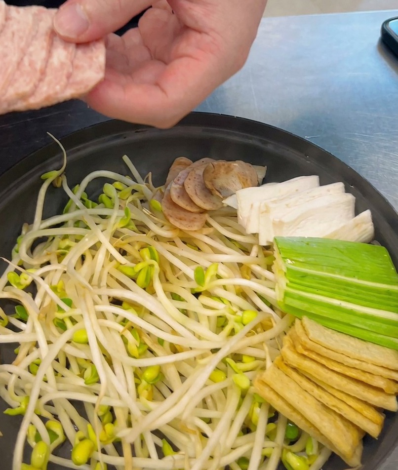 A persons hand placing uniform slices of pink spam into a shallow black pot already filled with bean sprouts and other stew ingredients.