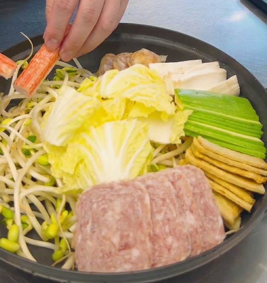 A hand arranging bright orange and white crab sticks into a pot already filled with sliced cabbage, spam, and tofu.