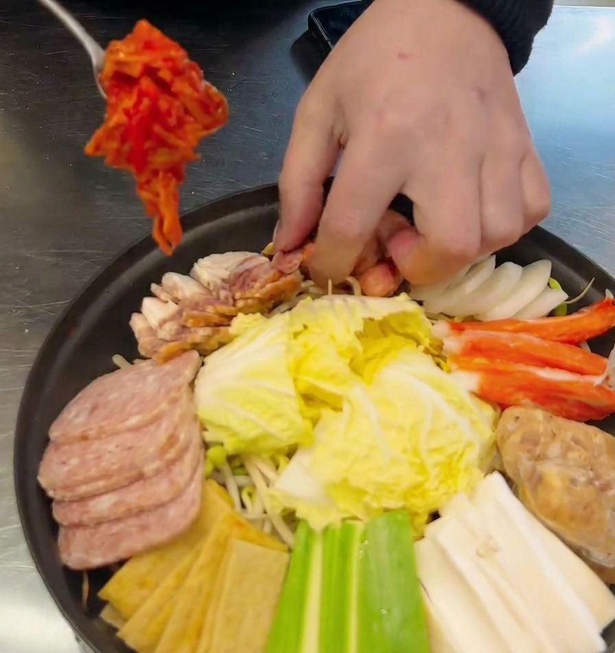 A hand using a metal spoon to place a portion of red kimchi into a shallow pot already filled with arranged sausages, spam, and vegetables.