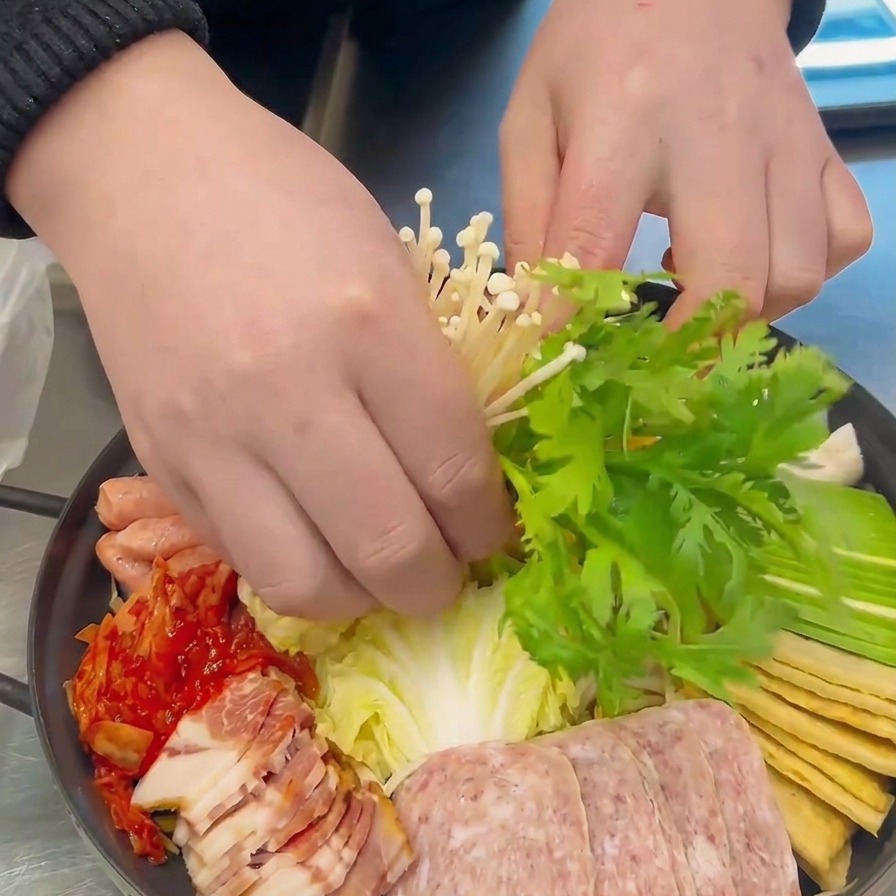 Hands carefully placing a cluster of white enoki mushrooms and green crown daisy leaves in the center of the prepared pot.