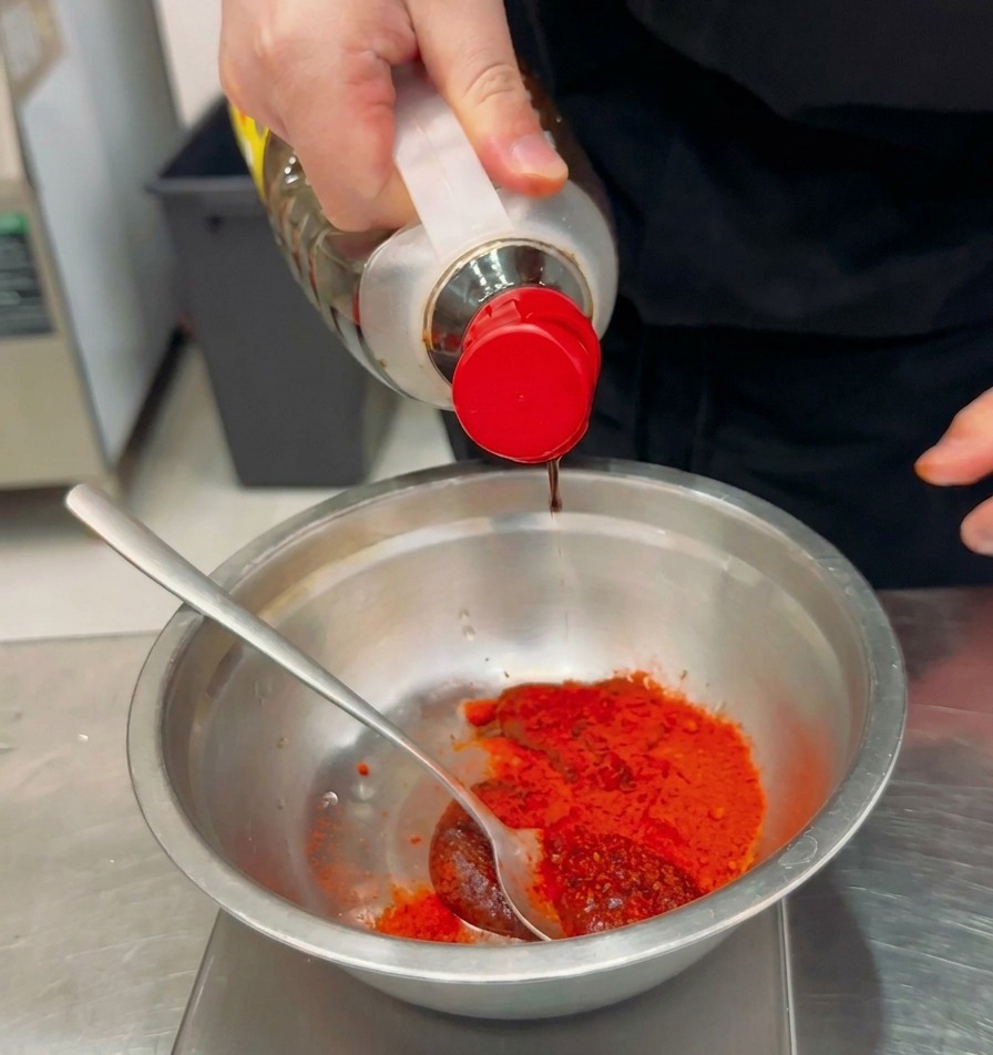 Liquid seasoning being poured from a clear bottle into a metal bowl containing red chili powder and paste.