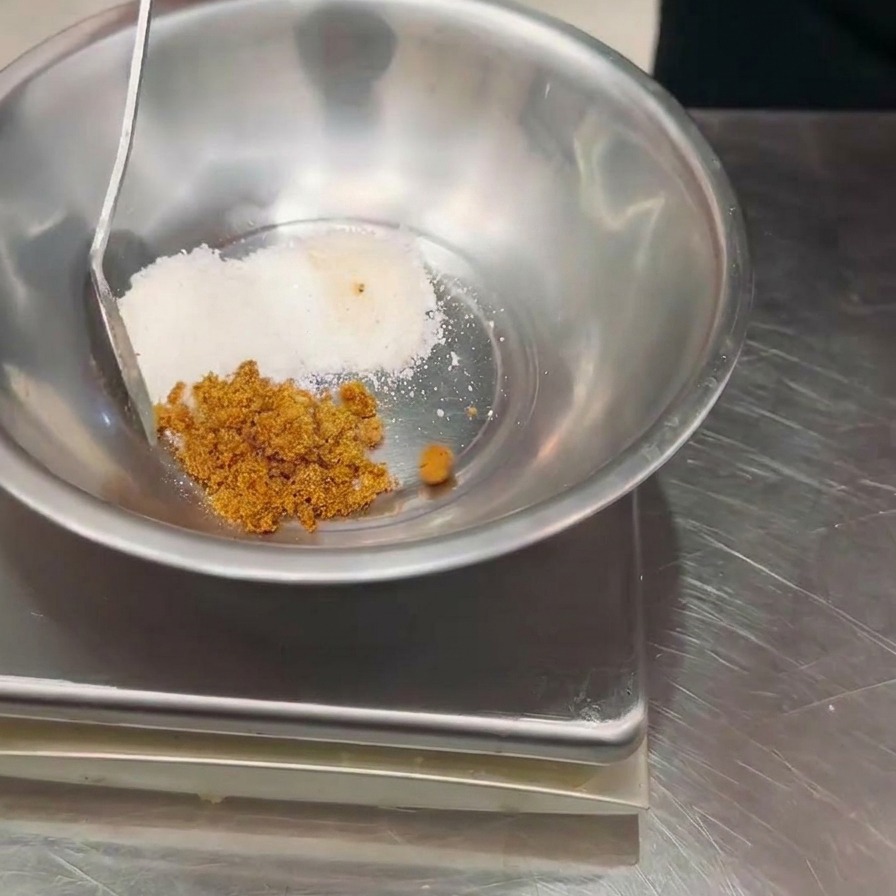 A hand using a spoon to add brown seasoning powder to a metal bowl containing white powder, placed on a digital scale.