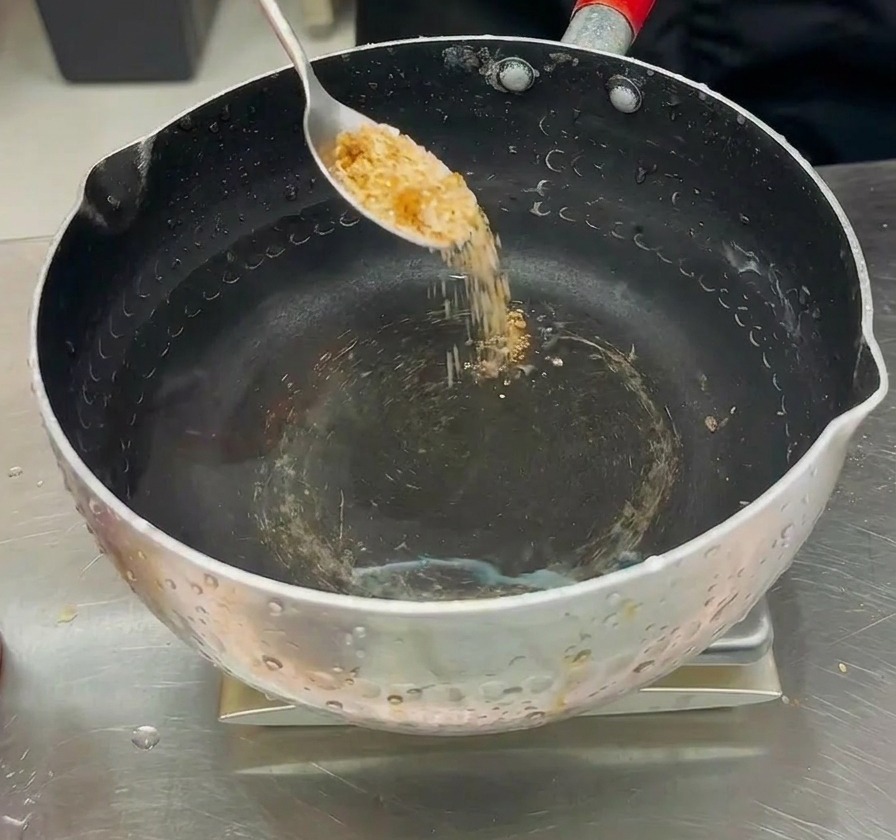 Seasoning powder being poured from a spoon into a pot of clear water to prepare the broth for the army stew.