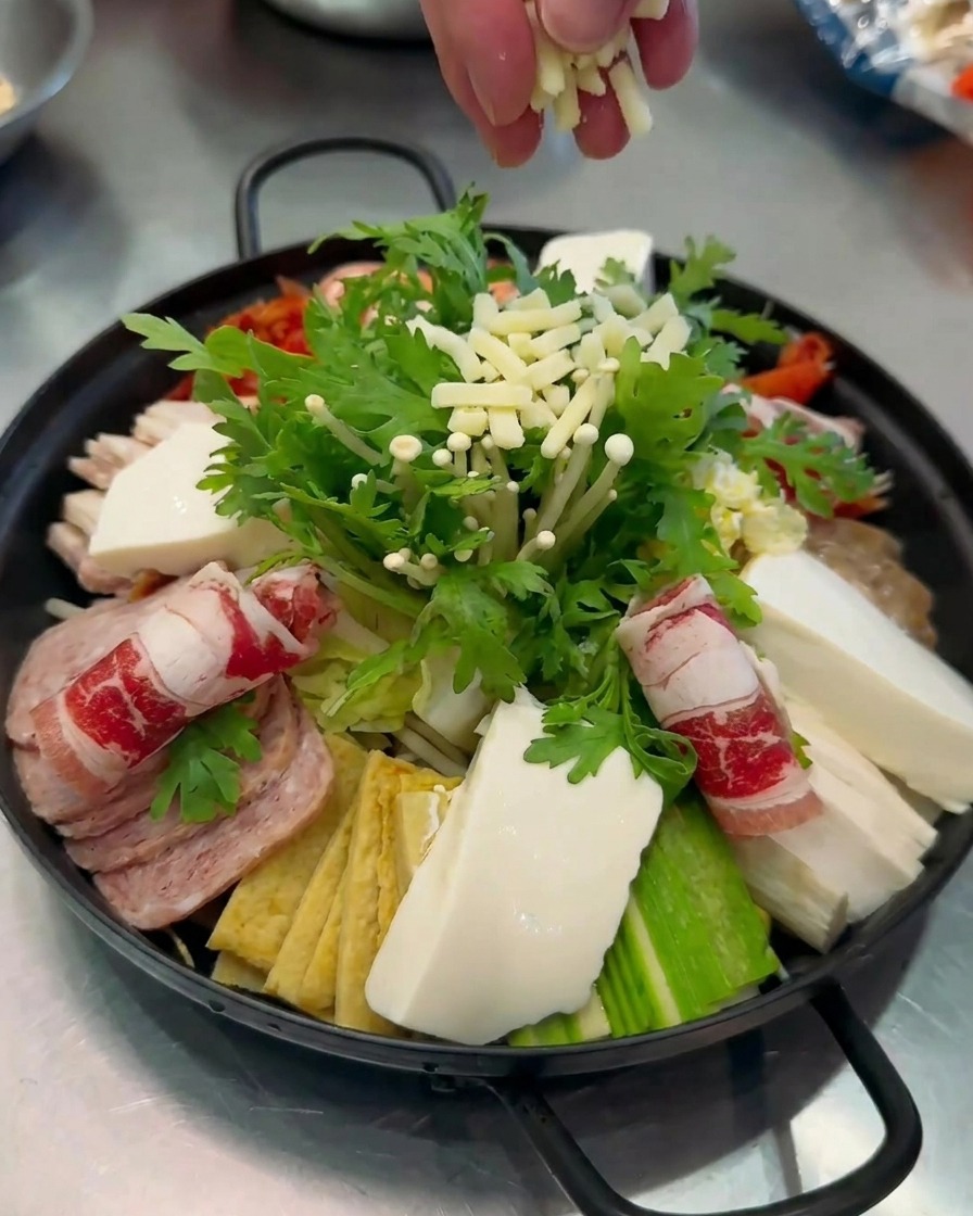 Close-up of shredded white cheese being sprinkled by hand over a colorful and fully assembled pot of Budae-jjigae.
