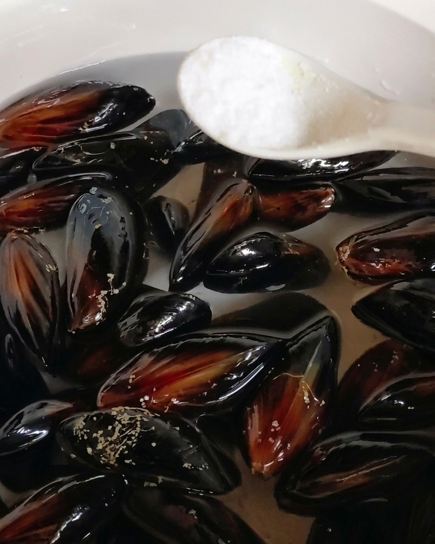 A spoonful of salt being added to a pot of black mussels submerged in water.