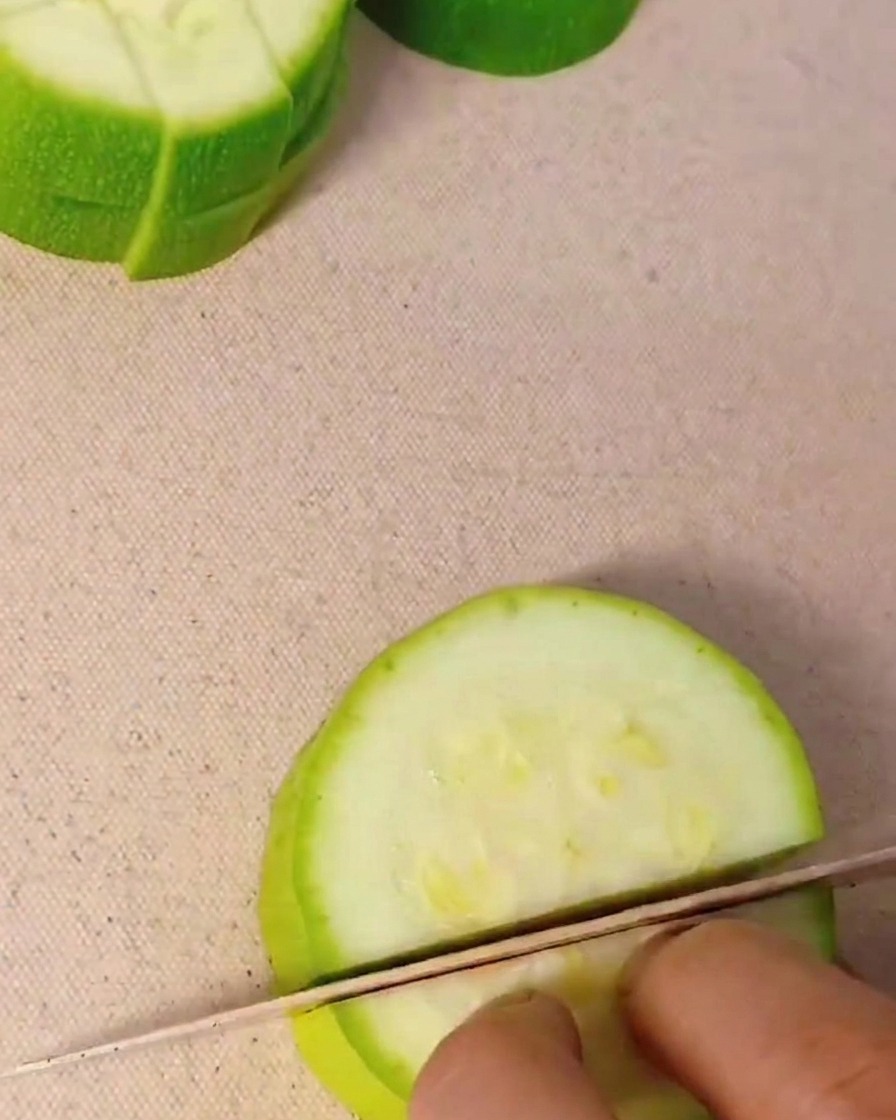 A close-up shot of a hand using a kitchen knife to cut a fresh green zucchini into small cubes on a light-colored cutting board.