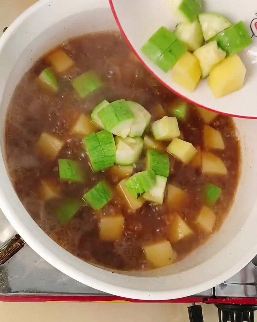 Diced green zucchini and yellow onions being poured from a small white plate into a bubbling reddish-brown broth in a pot.