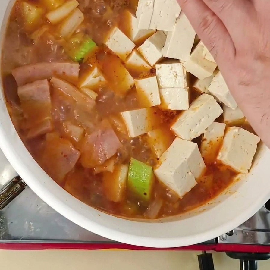 A hand placing several cubes of fresh white tofu into a pot of simmering Korean soybean paste stew containing zucchini and other ingredients.