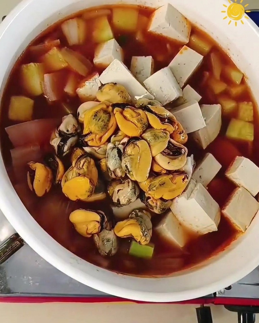 A cluster of orange mussel meat and several large grey raw prawns being added to the center of a boiling pot filled with tofu and vegetables.