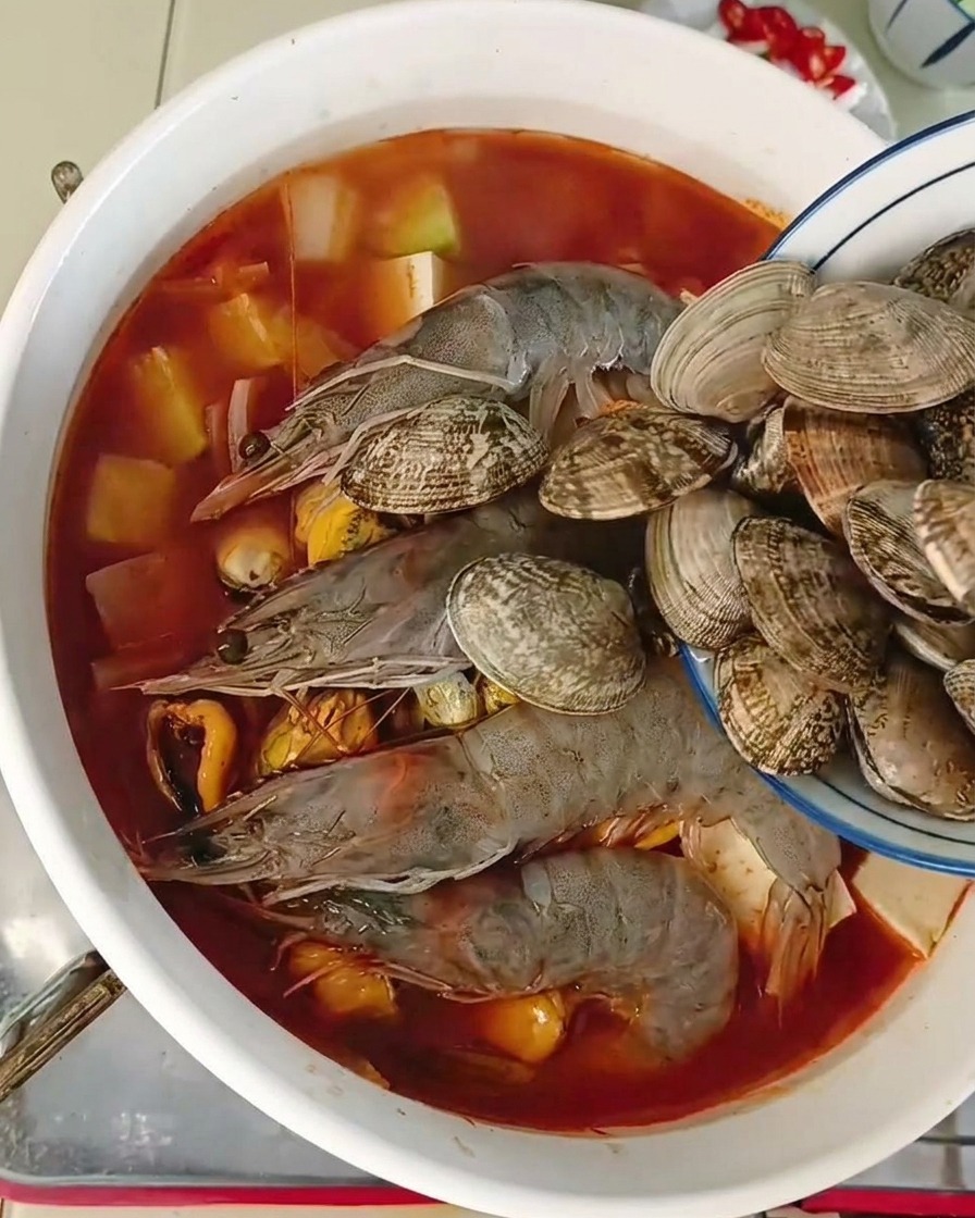 A small blue-rimmed bowl pouring fresh whole clams into a pot already filled with prawns mussels and tofu in a red-tinted broth