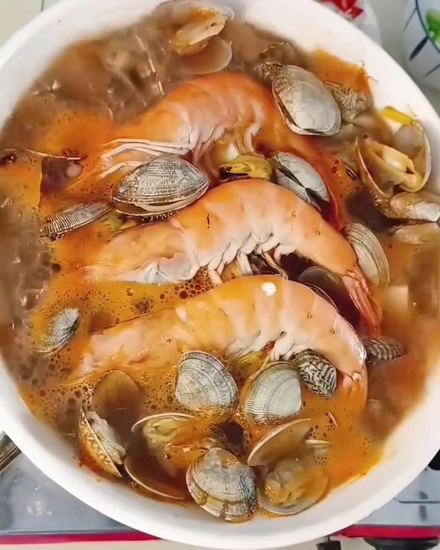 A close-up shot of a bubbling seafood stew in a white pot containing prawns, clams, and tofu, with fresh green chilies and scallions scattered on the surface.
