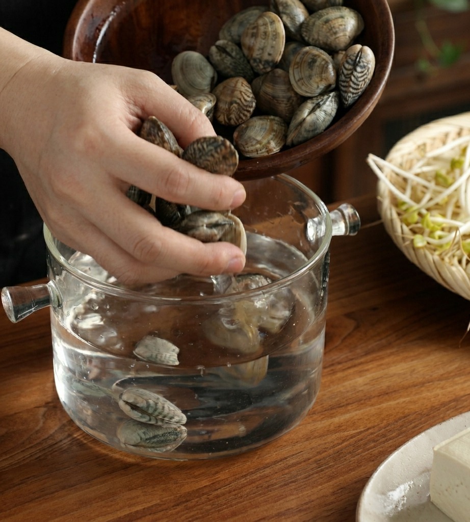 A hand pouring a bowl of fresh clams into a clear glass pot filled with water on a wooden table.