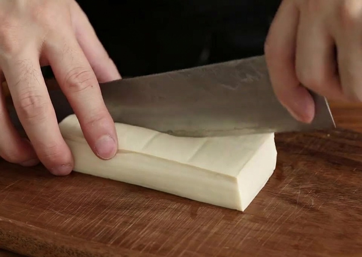 A hand using a large chef knife to slice a rectangular block of white tofu into smaller cubes on a dark wood board.