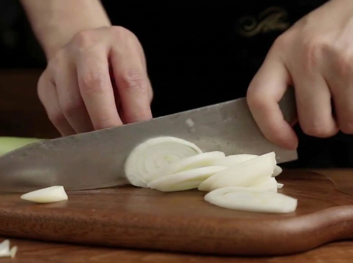 A close-up of hands using a knife to cut white leek rounds on a wooden cutting board.