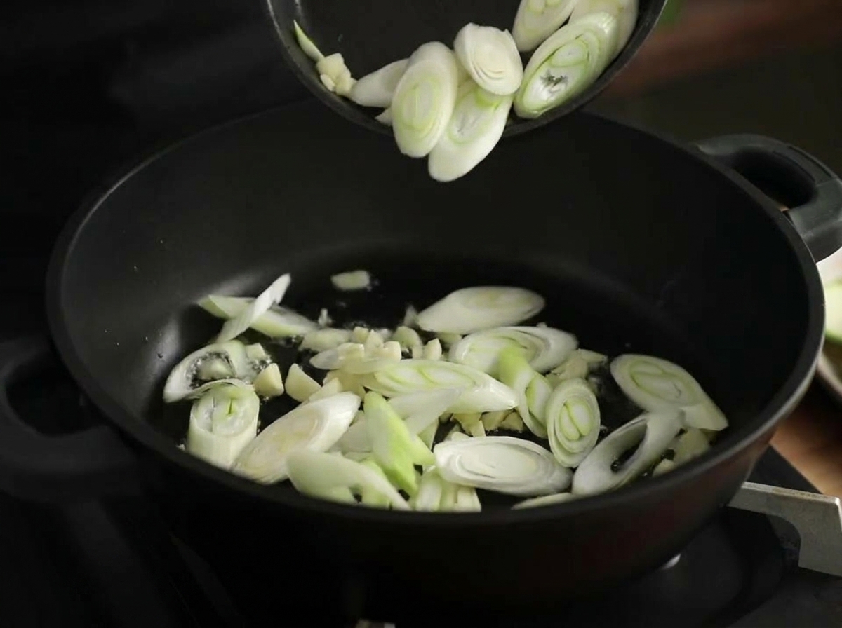 Minced garlic and diagonal slices of leek being stir-fried in a hot black pot with shimmering oil.