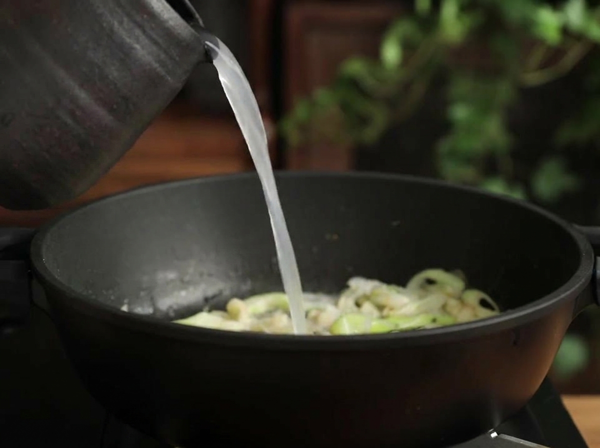 Cloudy white rice water being poured from a dark jug into a pot containing sautéed leeks and garlic.