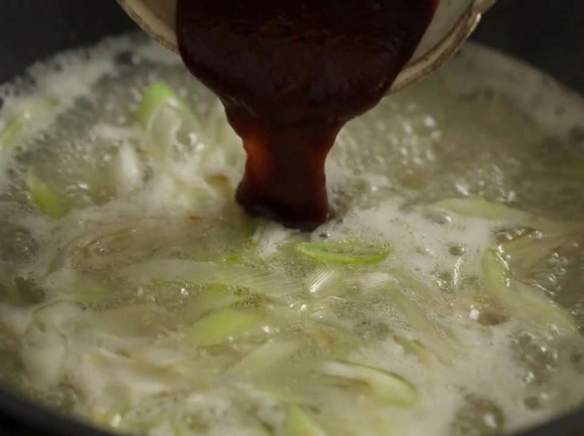 A large spoonful of dark red chili paste being stirred into a boiling pot of broth with leeks.