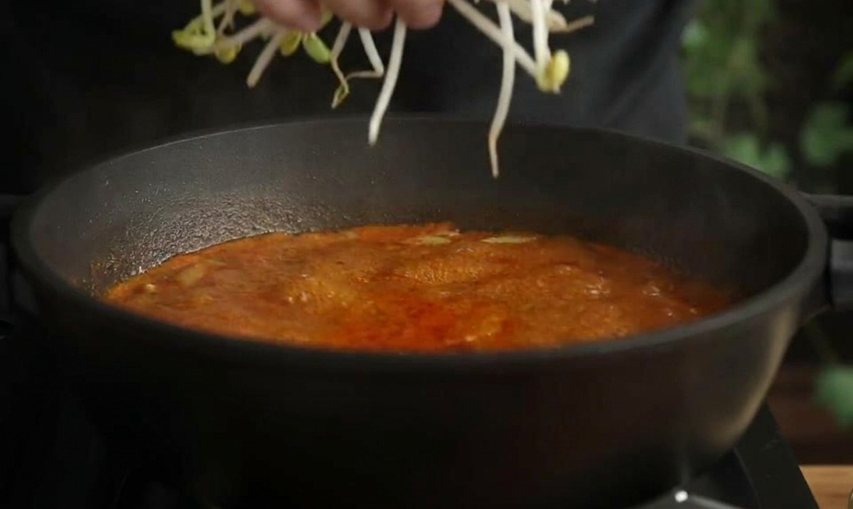 A hand adding a handful of long, pale bean sprouts into a boiling pot of red seafood soup.