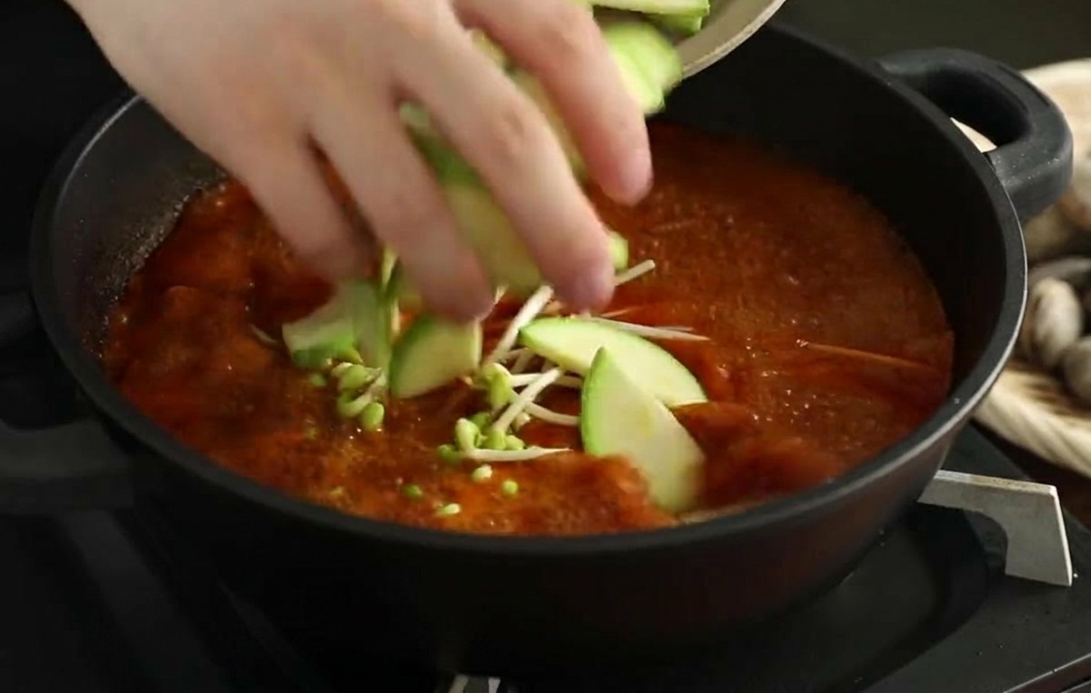 Hands placing fresh zucchini slices into a simmering red seafood broth that already contains bean sprouts.