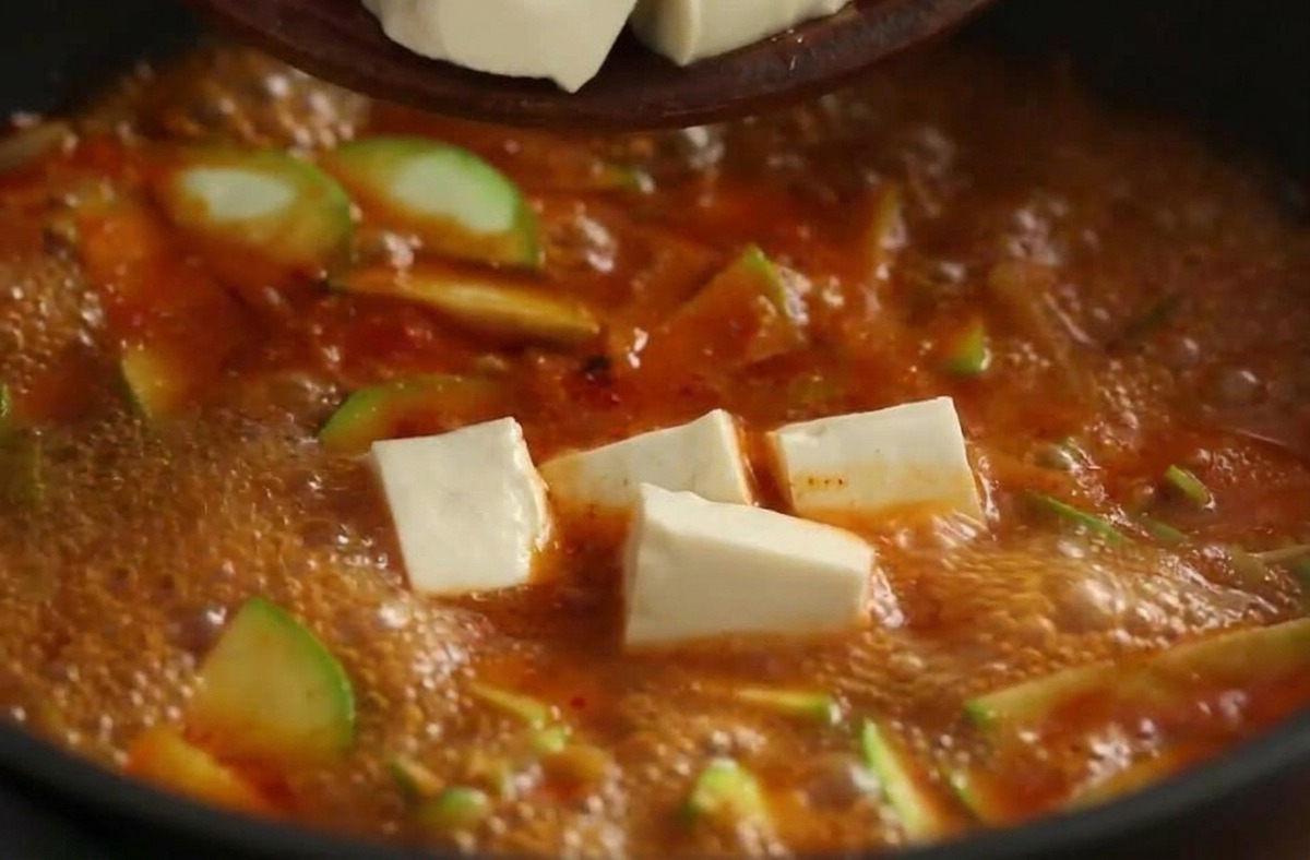 White cubes of tofu being dropped from a bowl into a vibrant, bubbling red Korean stew.