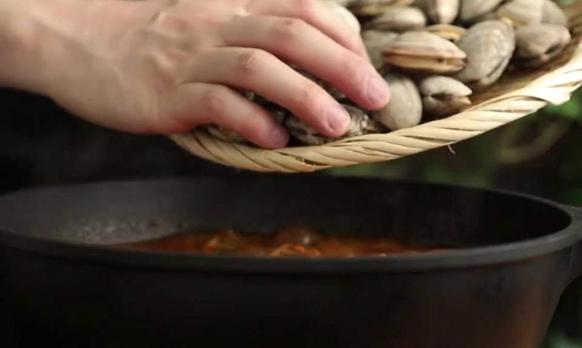 A person using a small bamboo strainer to lower fresh clams into a dark pot of simmering red stew.