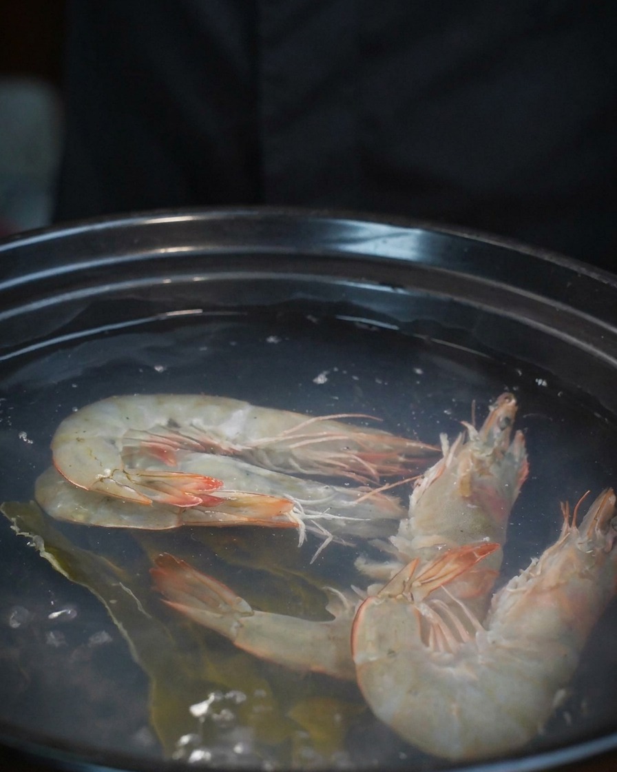 Fresh flower clams being poured from a bowl into a pot already containing shrimp and stock.