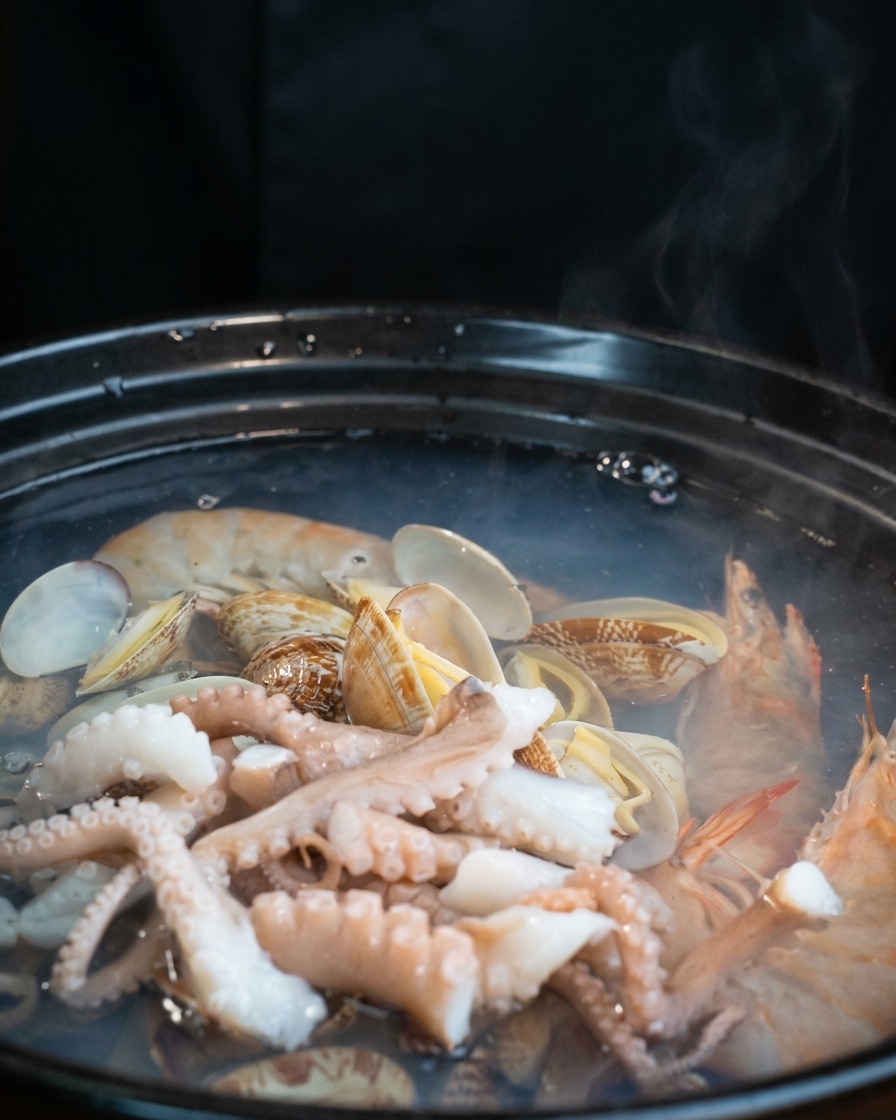 Slices of white tofu being layered on top of octopus and clams in the simmering pot.