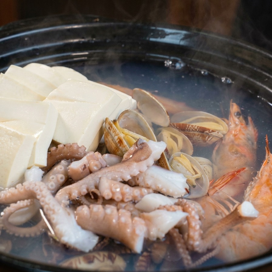 A simmering clay pot containing clams and shrimp with slices of white radish and orange carrots being added using a blue spatula.