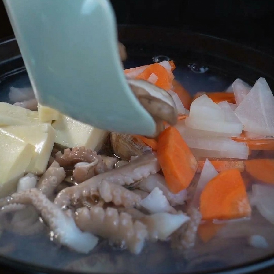 Slices of fresh shiitake mushrooms being added to a bubbling seafood soup alongside tofu, radish, and carrots.