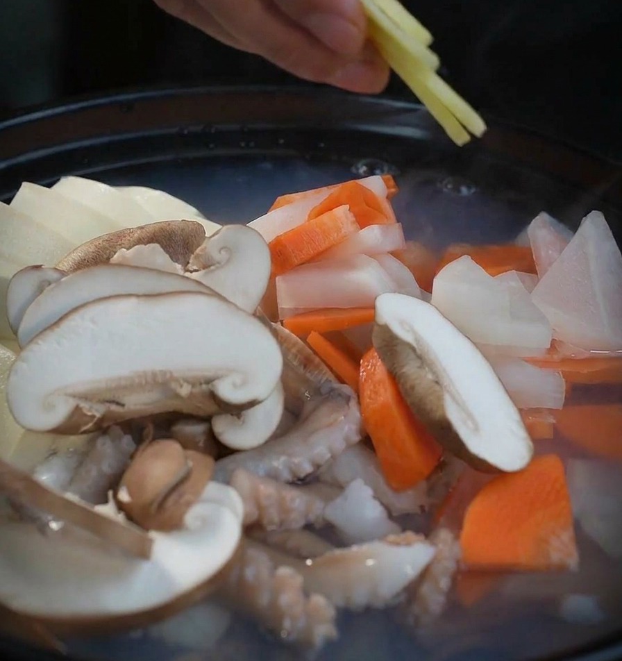 A hand placing thin slices of fresh ginger into a boiling pot filled with shiitake mushrooms, radish, and seafood.