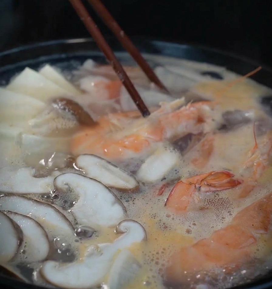 A steaming black pot filled with shrimp, mushrooms, and tofu simmering in a light broth, being stirred with dark wooden chopsticks.