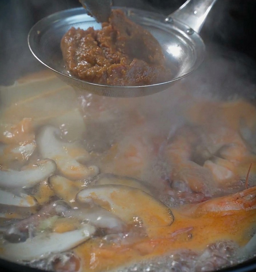 A scoop of thick, textured brown soybean paste held in a metal strainer over a bubbling pot of seafood soup.