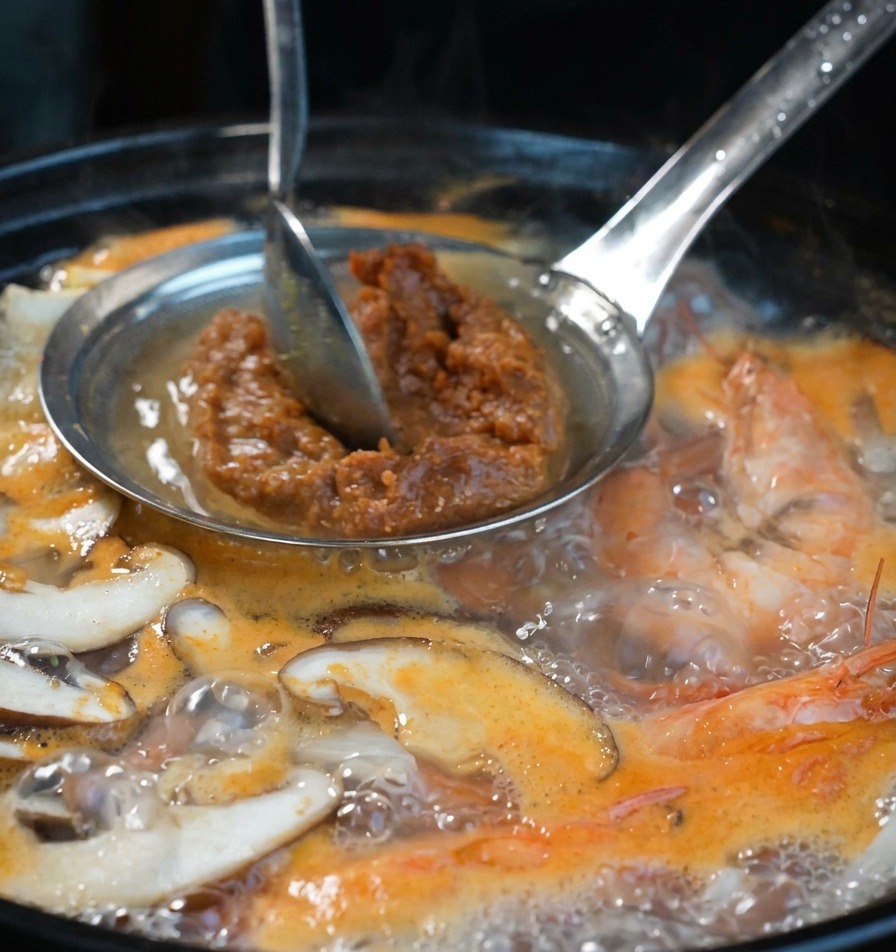 A hand using a metal spoon to press brown soybean paste through a strainer into a boiling seafood broth.