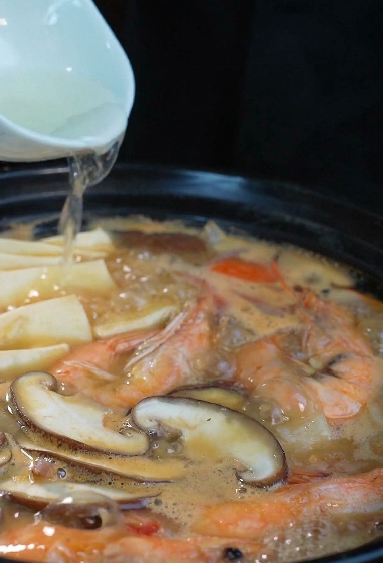 A stream of clear sake being poured from a white ceramic bowl into a steaming black pot filled with seafood and mushrooms.