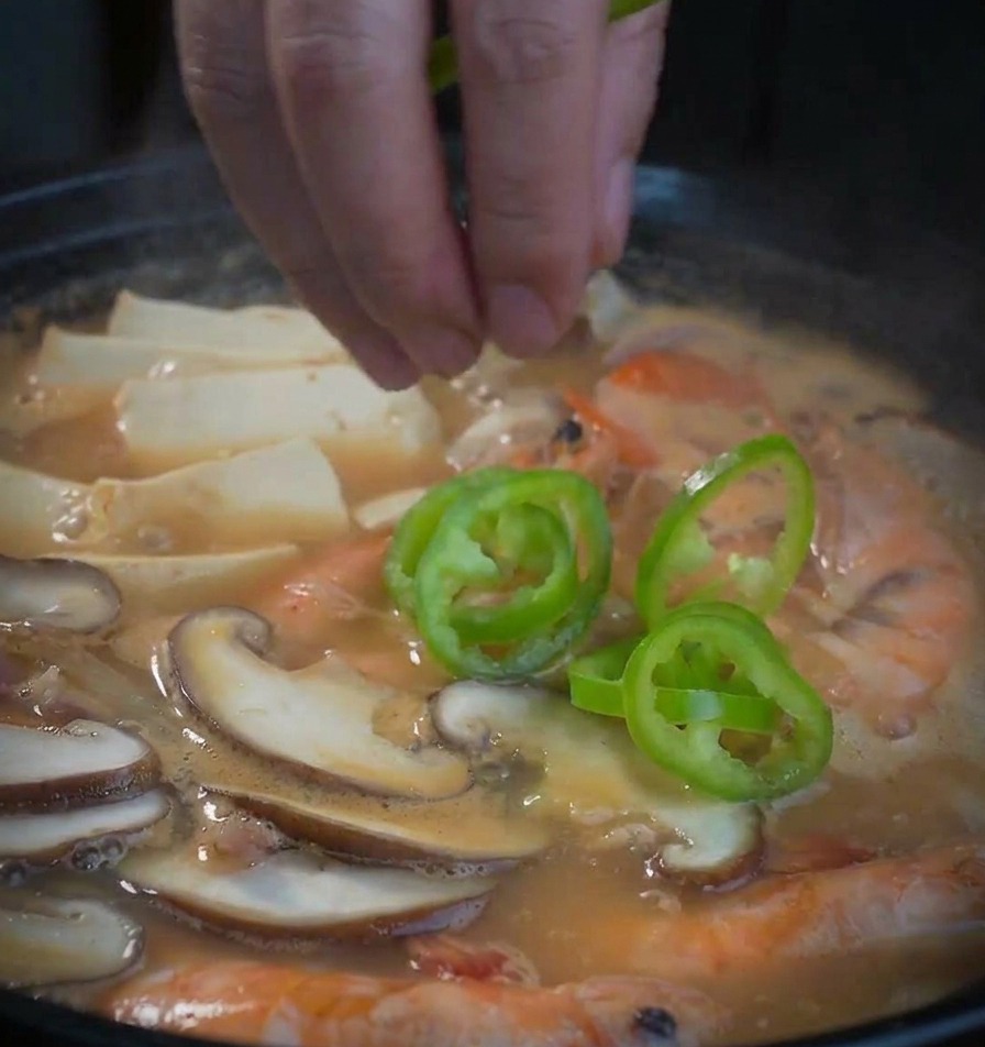 Green pepper rings being dropped by hand into a bubbling pot of seafood soybean paste soup.