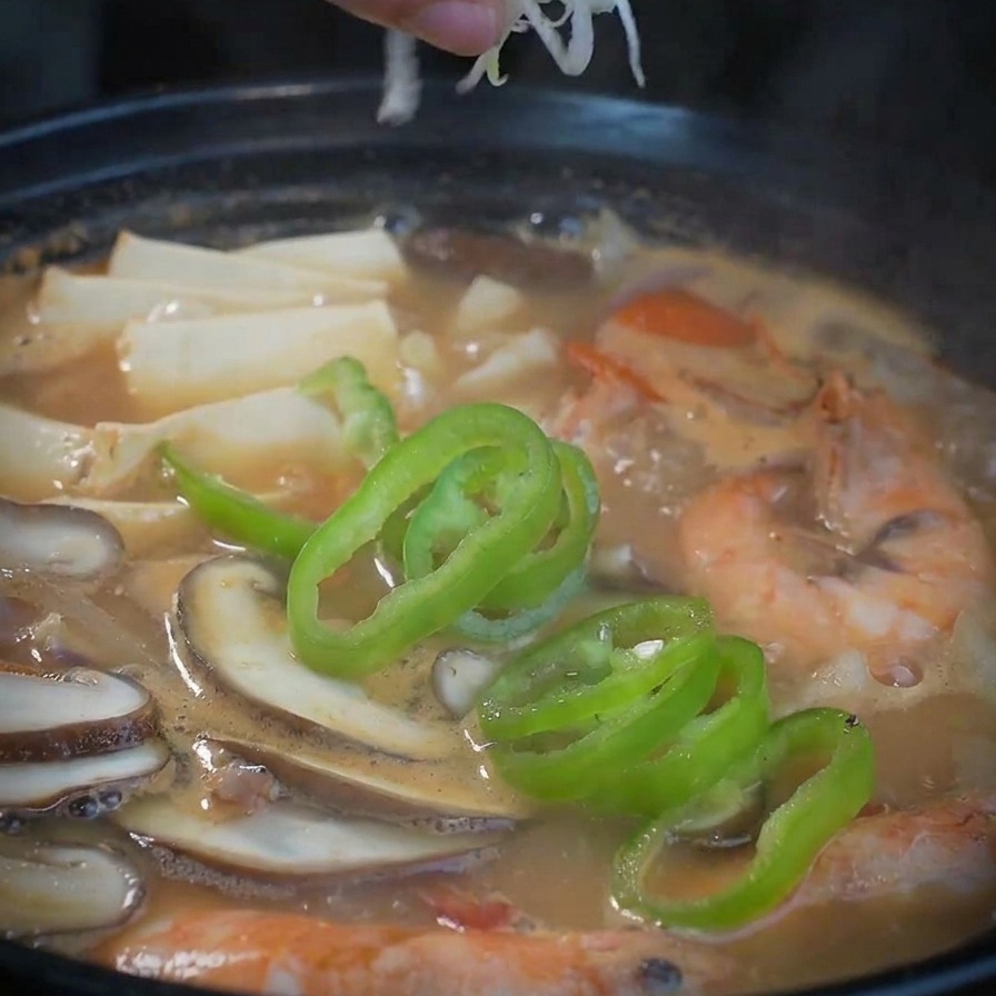 A hand scattering a mix of sliced white and green onions over a steaming pot of finished seafood soup.