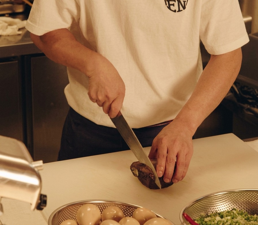 A chef slicing a slab of dark, braised pork belly into thick slices on a white cutting board.