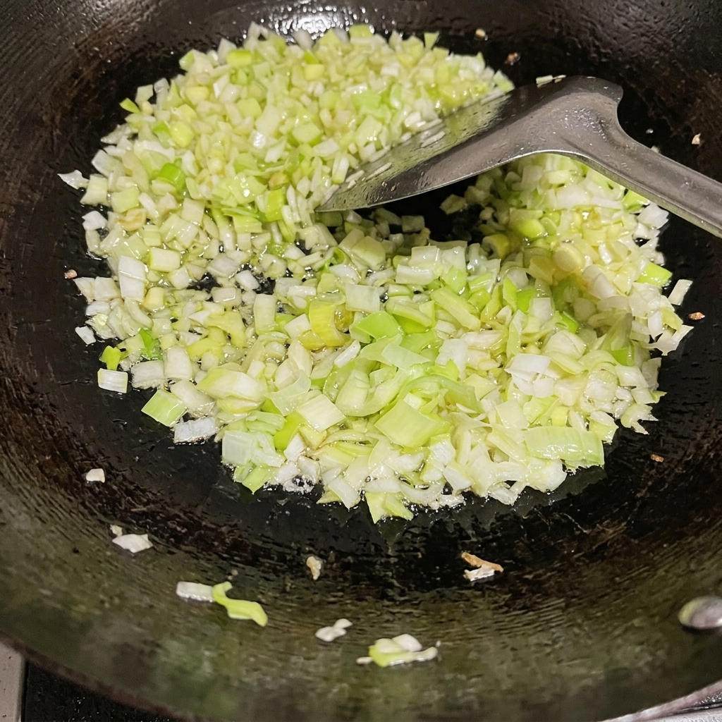Finely chopped green onions being stir-fried in a seasoned black wok with oil and a metal spatula.