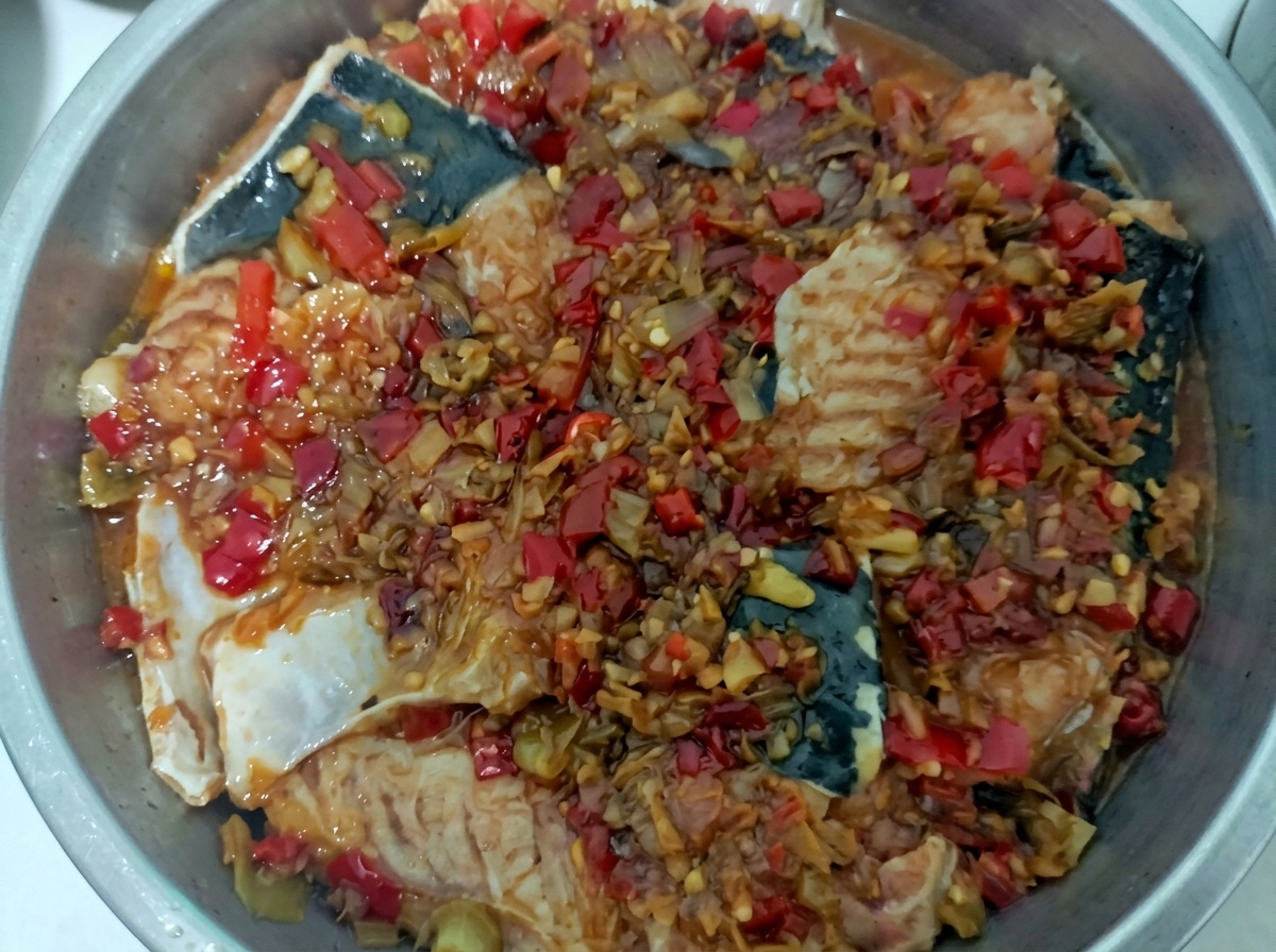 A stainless steel steaming tray filled with white fish pieces topped with a thick, vibrant red chopped chili and garlic sauce.
