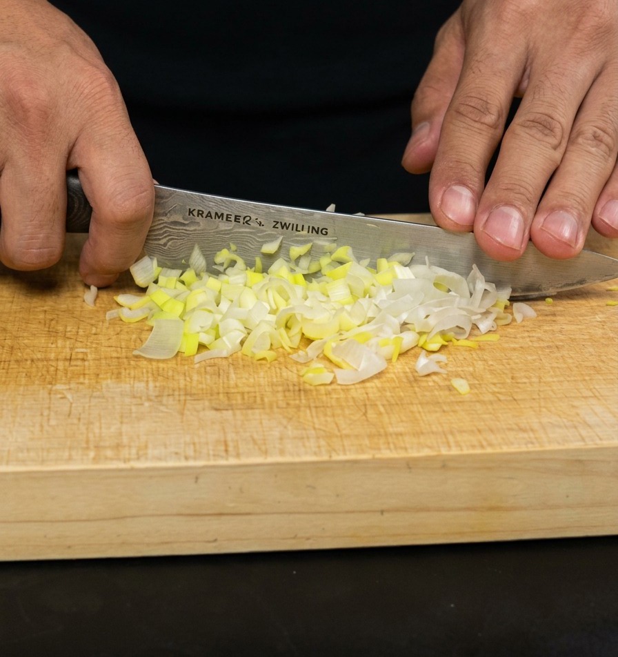 A persons hands using a chefs knife to finely mince green onions on a light-colored wooden cutting board.