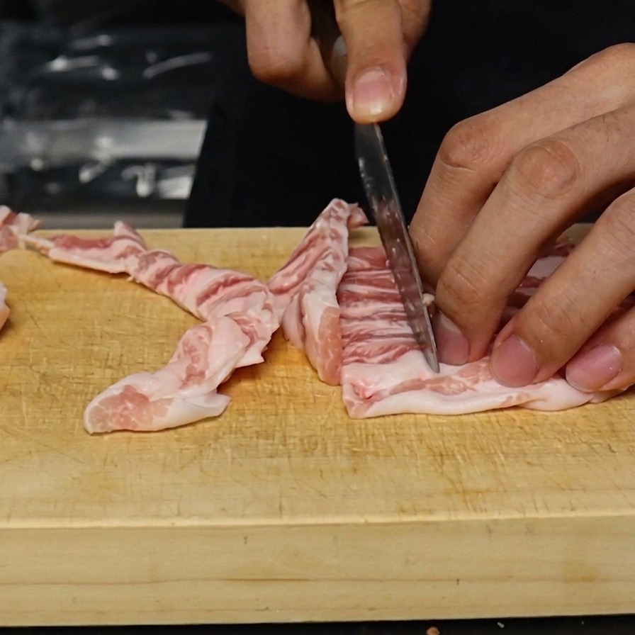 Raw pork belly being sliced into thin, even one-centimeter strips on a wooden cutting board.