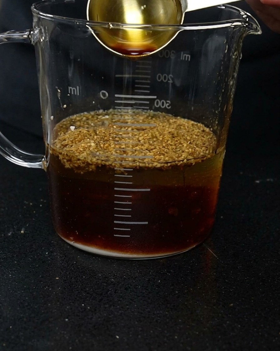 A gold measuring spoon pouring dark soy sauce into a glass measuring cup filled with water and various seasoning powders.
