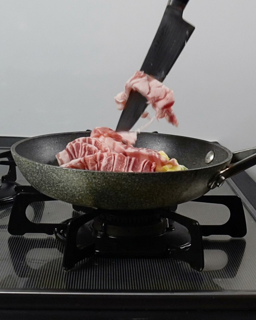 A knife pushing strips of raw pork belly into a pan containing chopped green onions and garlic.