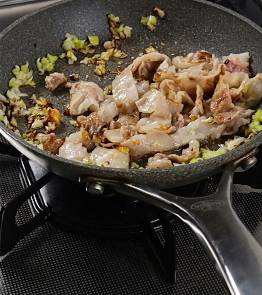 Strips of pork belly and minced green onions being stir-fried in a speckled pan until the meat surface appears browned.