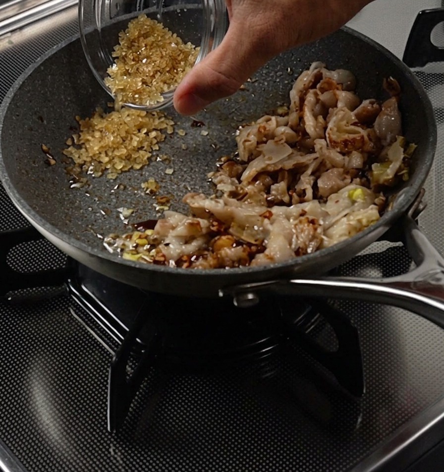 A hand pouring brown sugar from a small bowl into a cleared area of a frying pan next to cooked pork strips.
